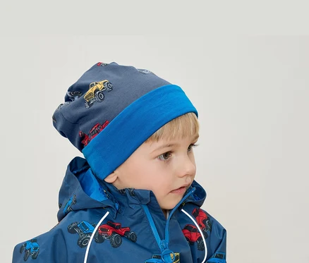Un garçon blond porte un bonnet bleu et une veste avec un imprimé de monster trucks.
