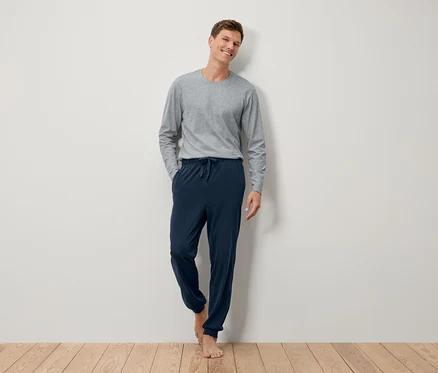 Un homme pieds nus se tient contre un mur, portant un T-shirt gris à manches longues et un pantalon de survêtement bleu marine.