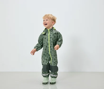 Un garçon en combinaison verte à motif de taches et bottes en caoutchouc vertes saute dans une flaque d'eau.