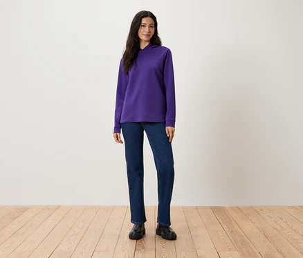 Une femme debout portant un sweat à capuche violet, un jean et des chaussures noires.