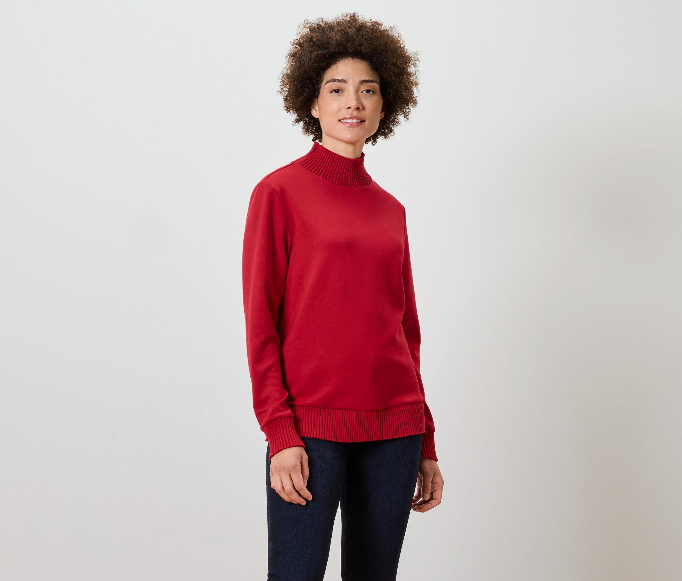 Une femme aux cheveux bouclés porte un pull à col roulé rouge et un jean bleu foncé.