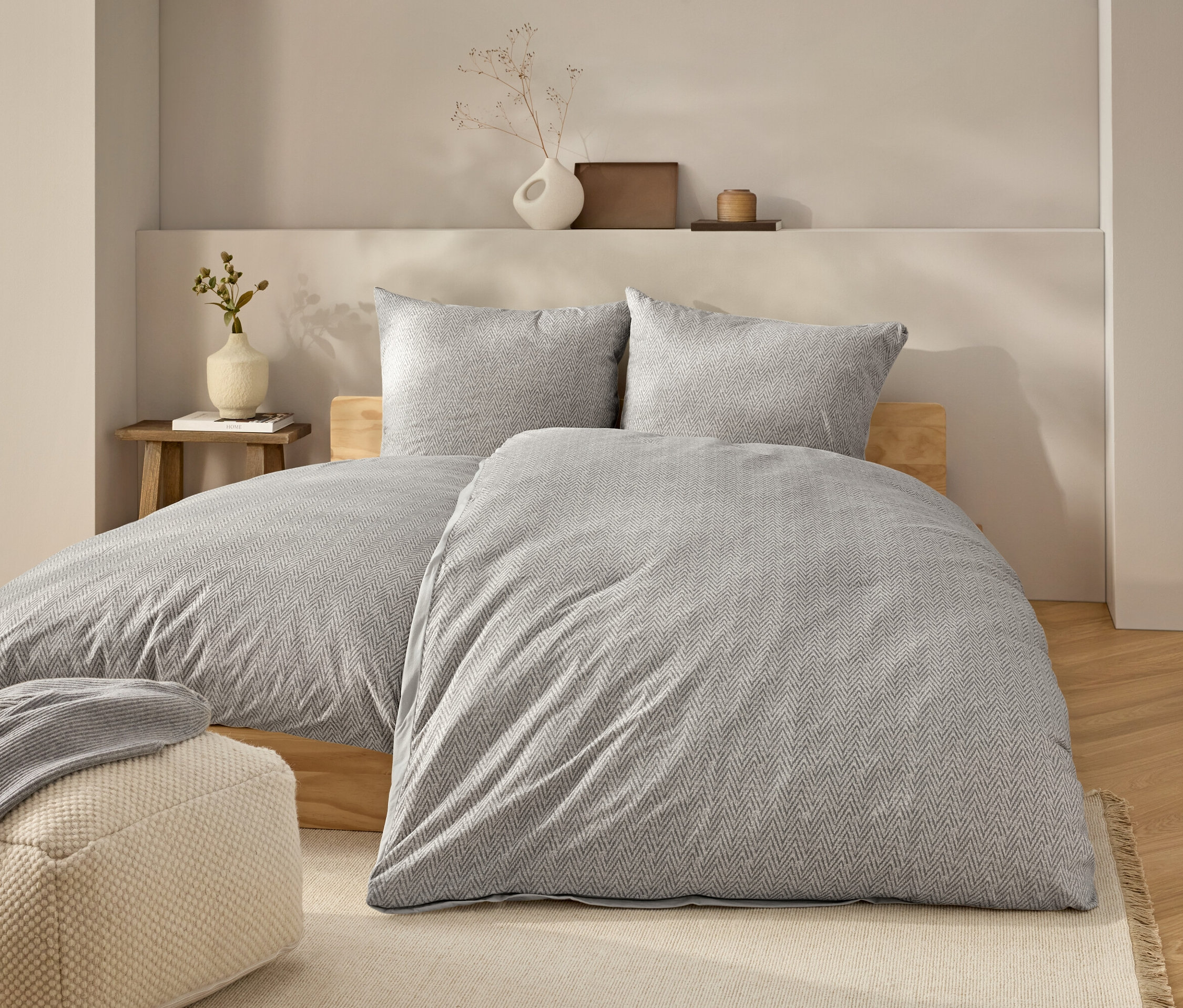Lit avec des draps gris et deux oreillers. Une couverture grise est posée sur le lit. Un pouf beige se trouve à côté du lit.