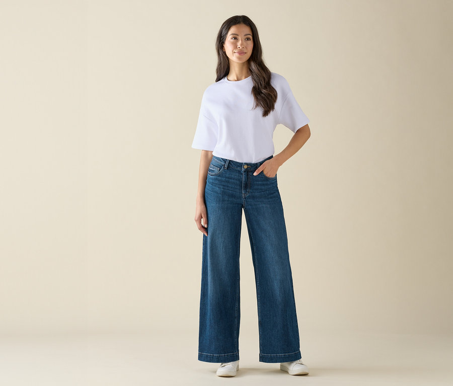 Une femme portant un t-shirt blanc et un jean bleu large pose sur un fond clair.