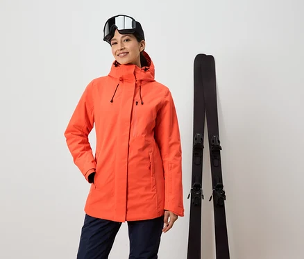 Femme en veste orange et lunettes de ski debout à côté des skis.