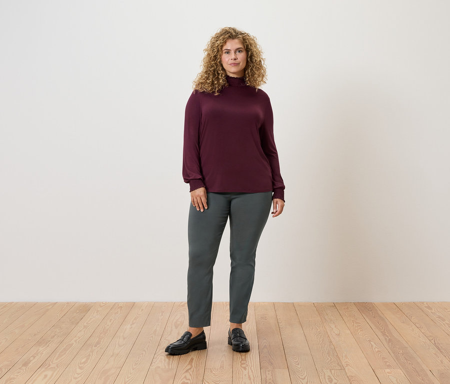 Une femme debout sur un plancher en bois porte un pantalon stretch vert foncé et un haut à manches longues bordeaux.