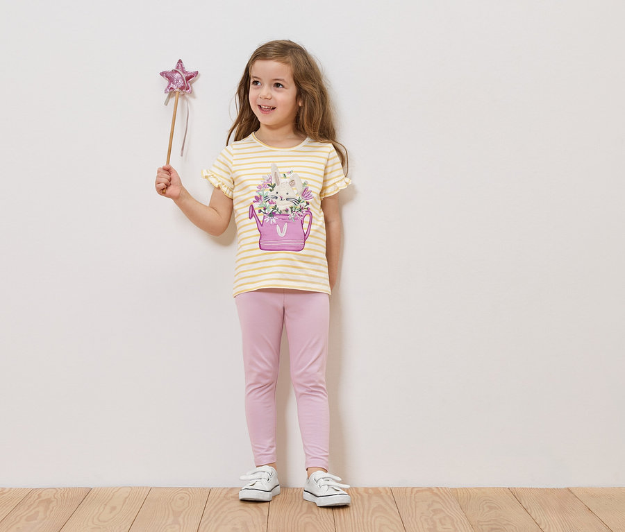 Une fille tient une baguette étoilée, elle porte un t-shirt rayé avec un arrosoir et des fleurs, ainsi qu'un legging rose.