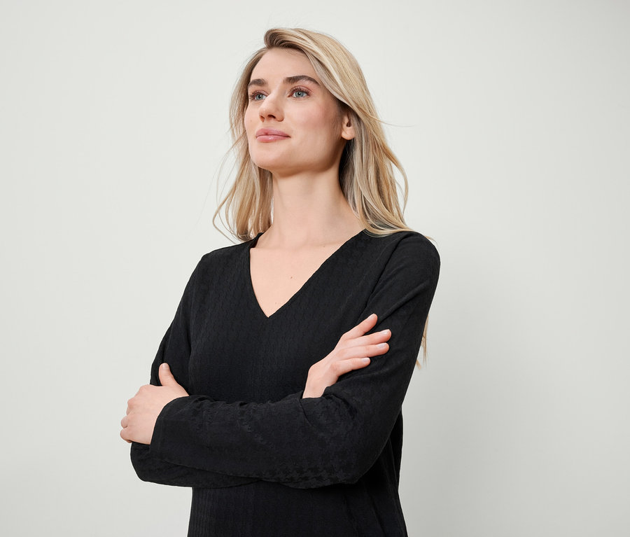 Une femme blonde, les bras croisés, porte un t-shirt noir à manches longues avec un col en V.