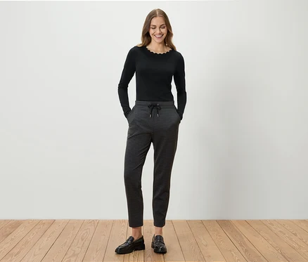 Une femme se tient debout et sourit, elle porte un haut noir à manches longues, un pantalon gris à motifs et des mocassins noirs.