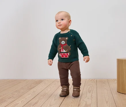 Un bébé vêtu d'un pull vert avec un ours jouant du tambour, d'un pantalon marron et de bottes se tient sur un plancher en bois.