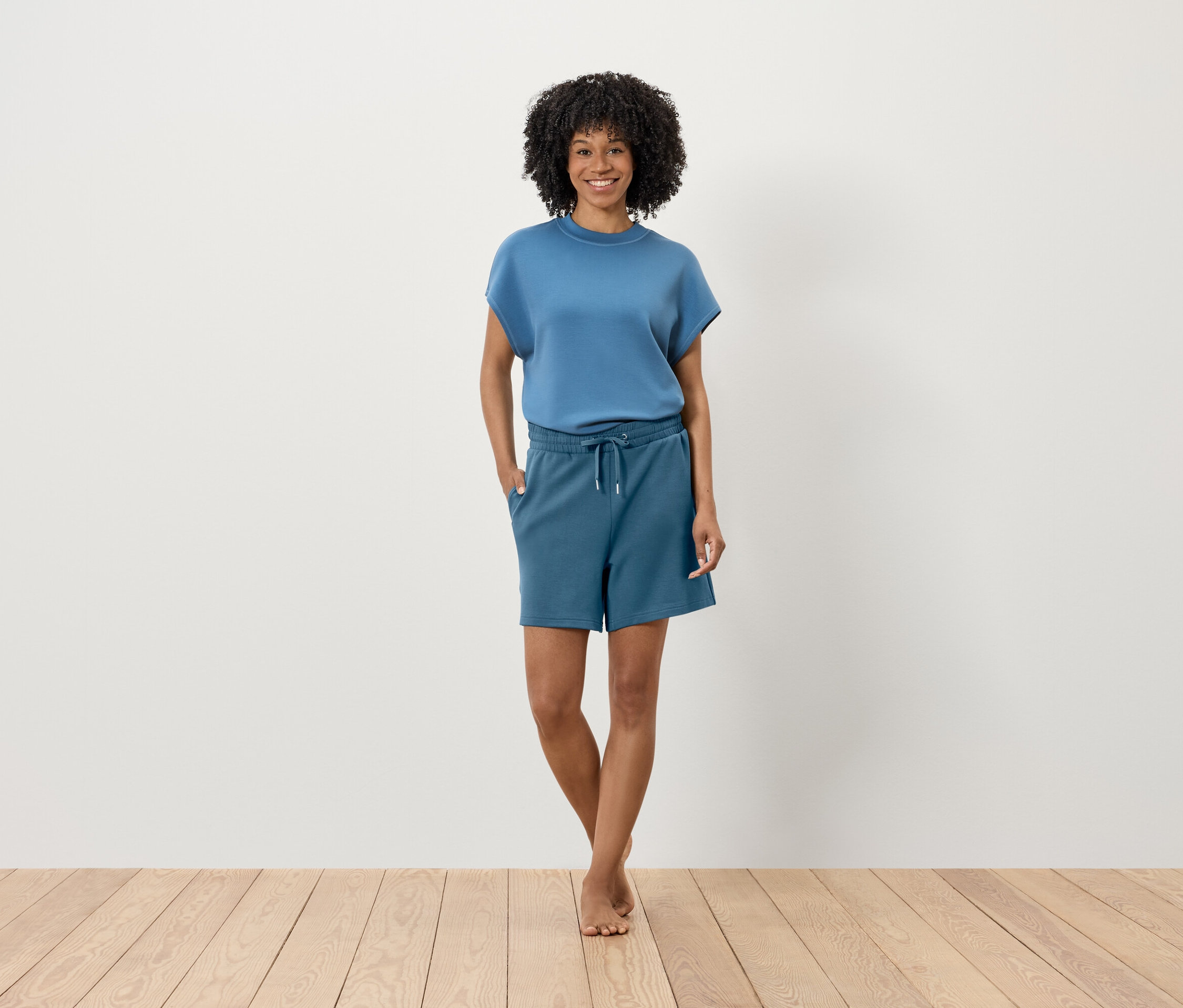 Une femme avec une coiffure afro se tient sur un plancher en bois devant un mur blanc. Elle porte un t-shirt bleu et un short.