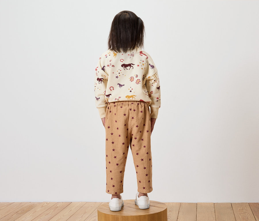 Une fille se tient dos à la caméra, portant un sweat-shirt crème avec un imprimé de chevaux et un pantalon marron avec des étoiles.