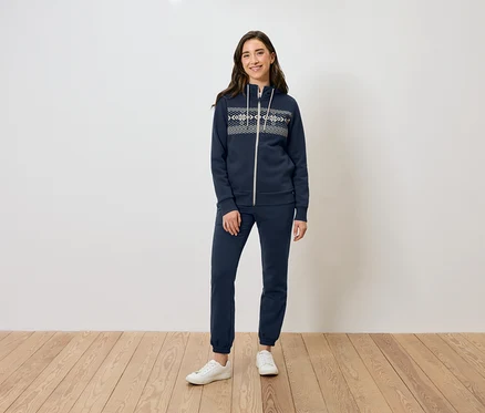 Une femme se tient sur un plancher en bois et porte un sweat à capuche zippé bleu marine, un pantalon de survêtement et des baskets blanches.