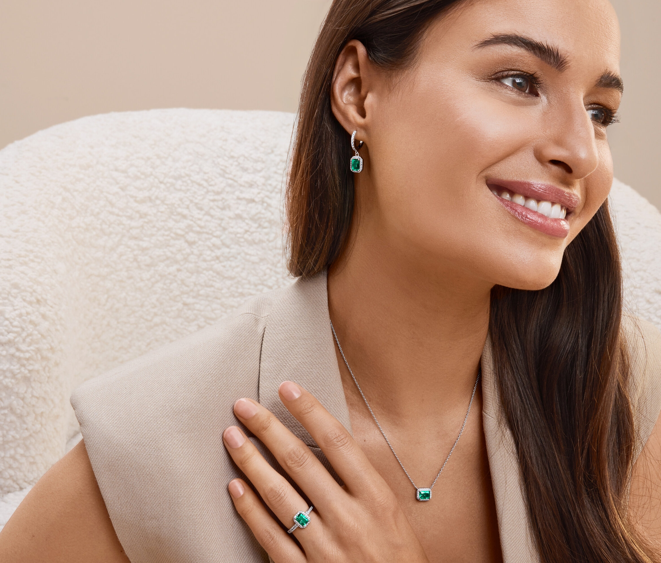 Une femme souriante est assise dans un fauteuil et porte des boucles d'oreilles, un collier et une bague avec des pierres vert émeraude.