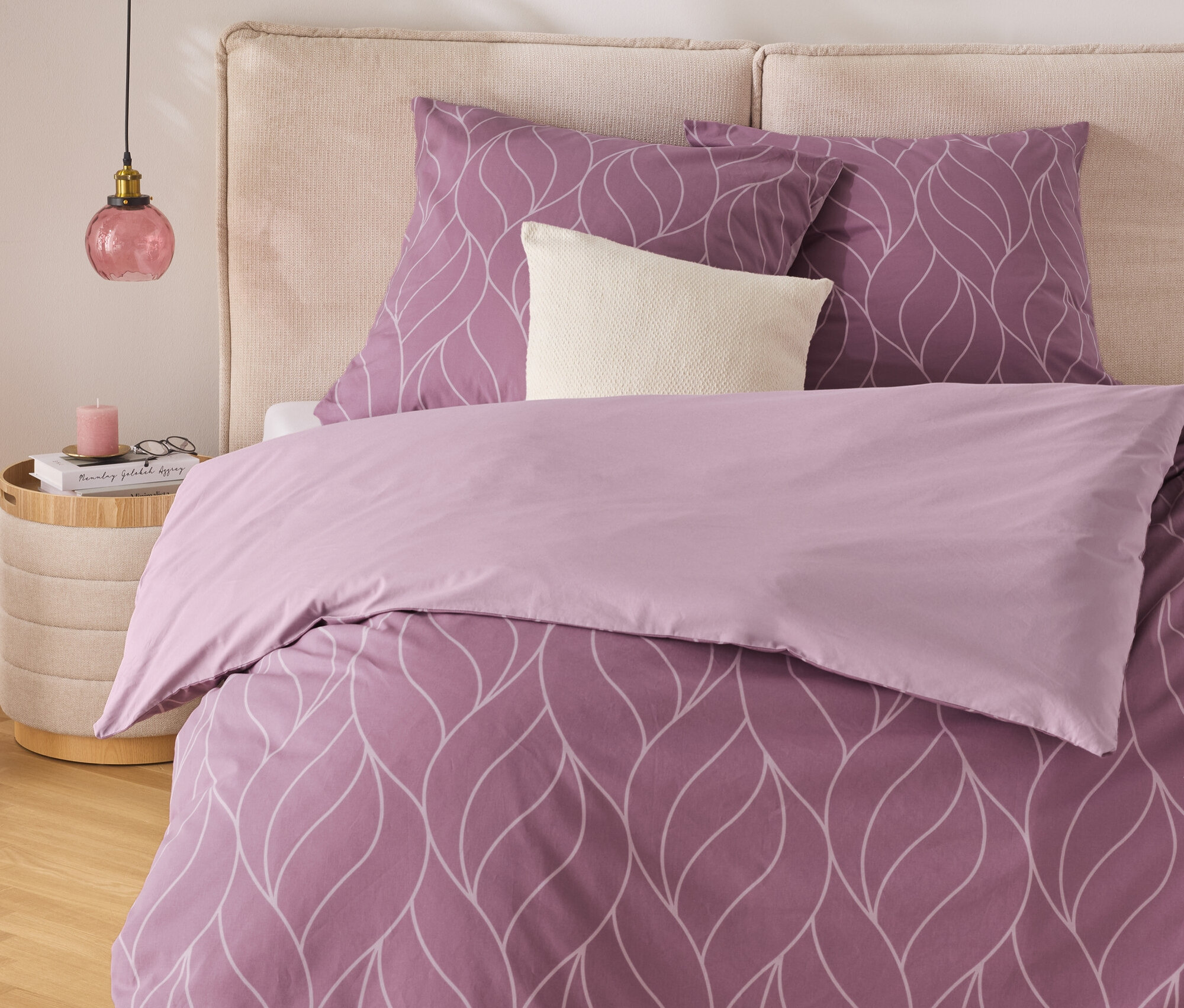 Linge de lit violet avec motif de feuilles blanches sur un lit avec tête de lit beige. Une table de chevet avec une lampe se trouve à côté du lit.