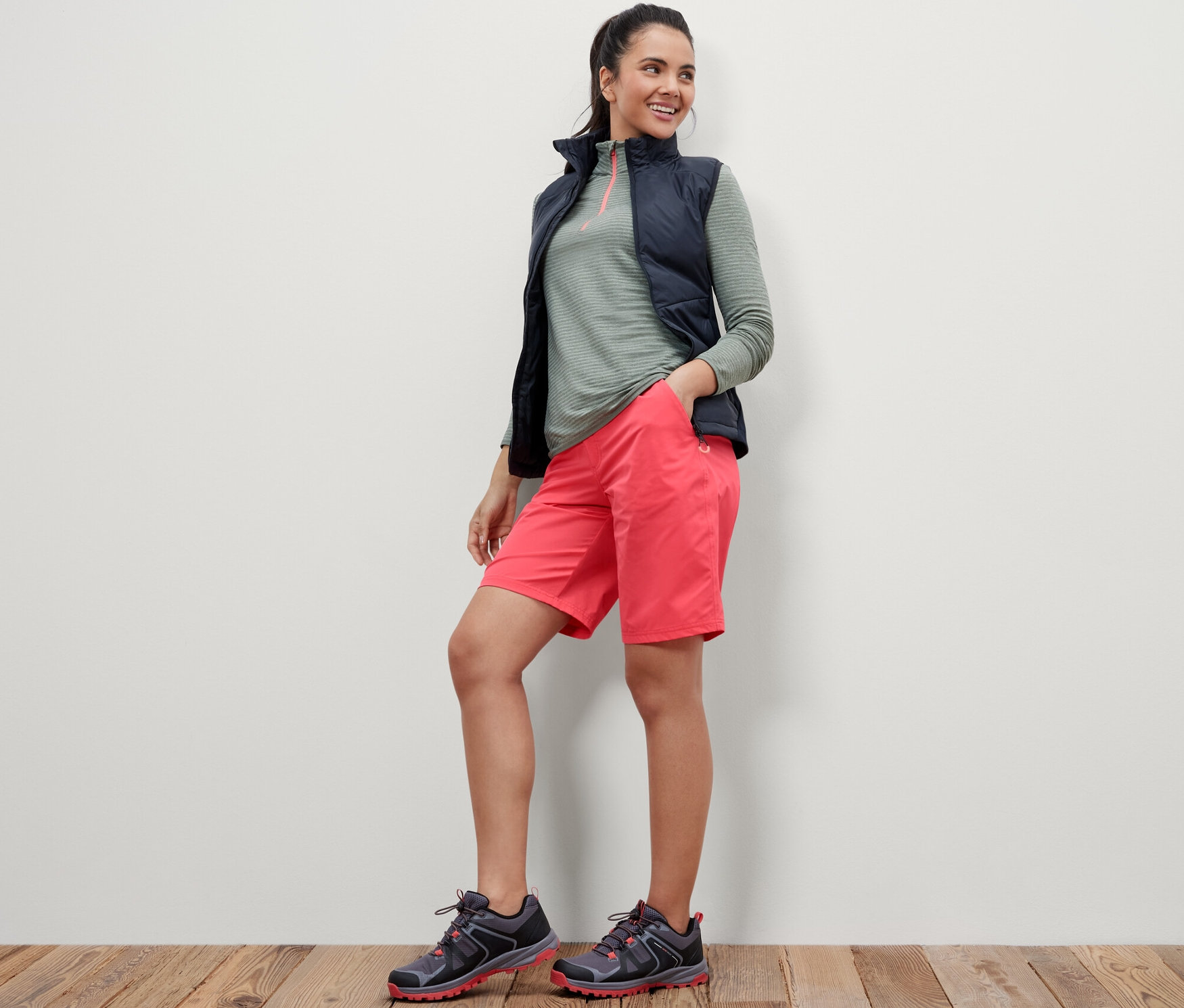 Une femme se tient debout, les mains dans les poches d'un short fonctionnel en softshell rose.