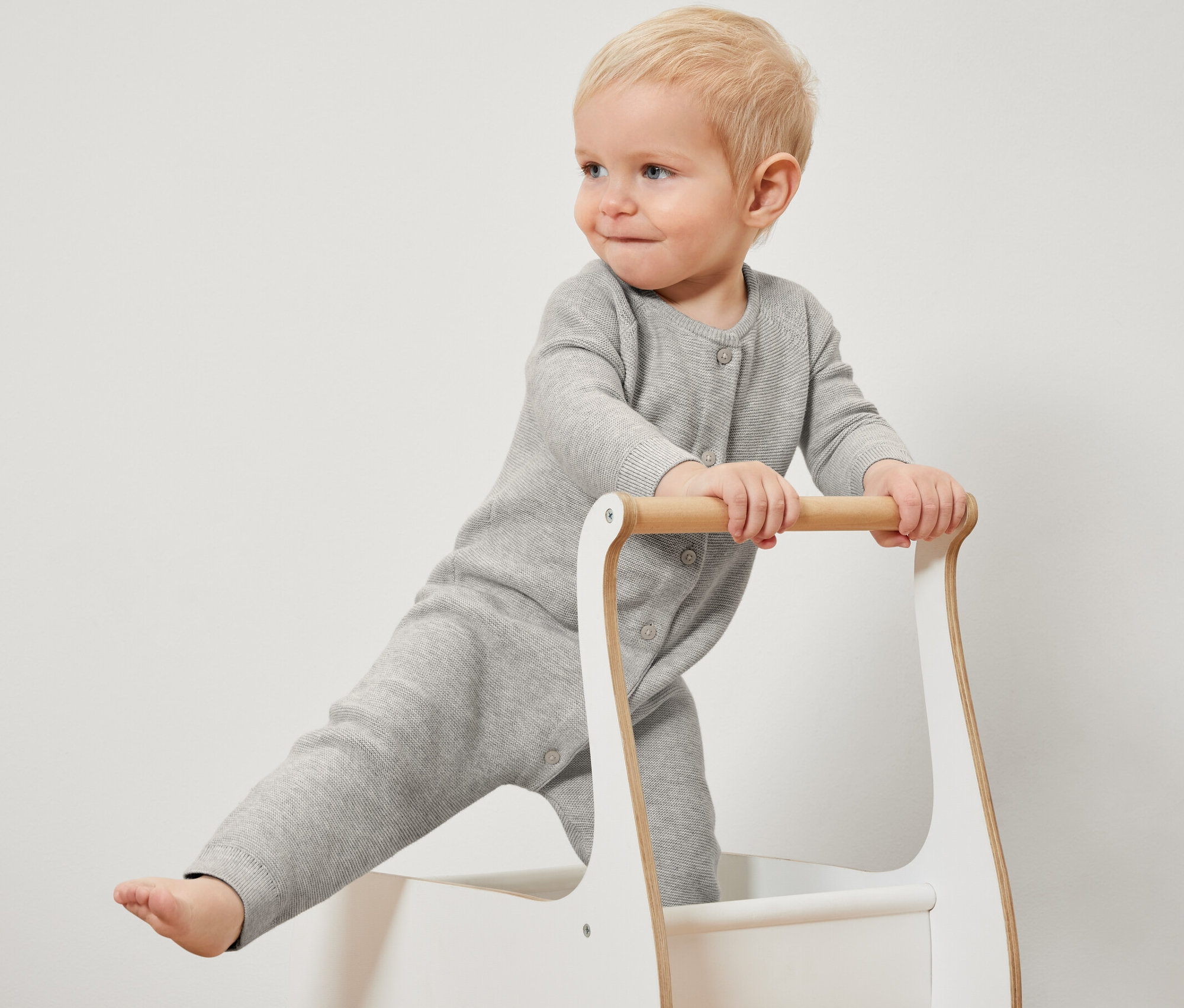 Un jeune enfant blond se tient dans un trotteur et porte une combinaison en maille grise.