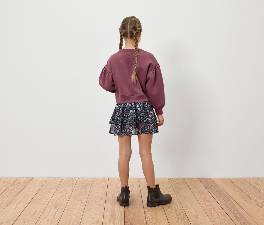 Une fille avec des tresses se tient dos à la caméra, portant un sweat-shirt bordeaux, une jupe à fleurs et des bottes marron.