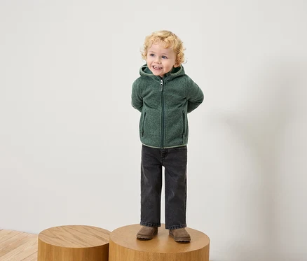 Un garçon aux cheveux blonds bouclés se tient sur un socle en bois, portant un sweat à capuche en polaire vert, un jean noir et des chaussures marron.
