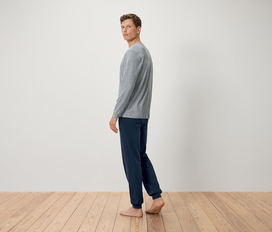 Un homme en t-shirt gris à manches longues et pantalon bleu marine se tient pieds nus sur un plancher en bois et regarde par-dessus son épaule.