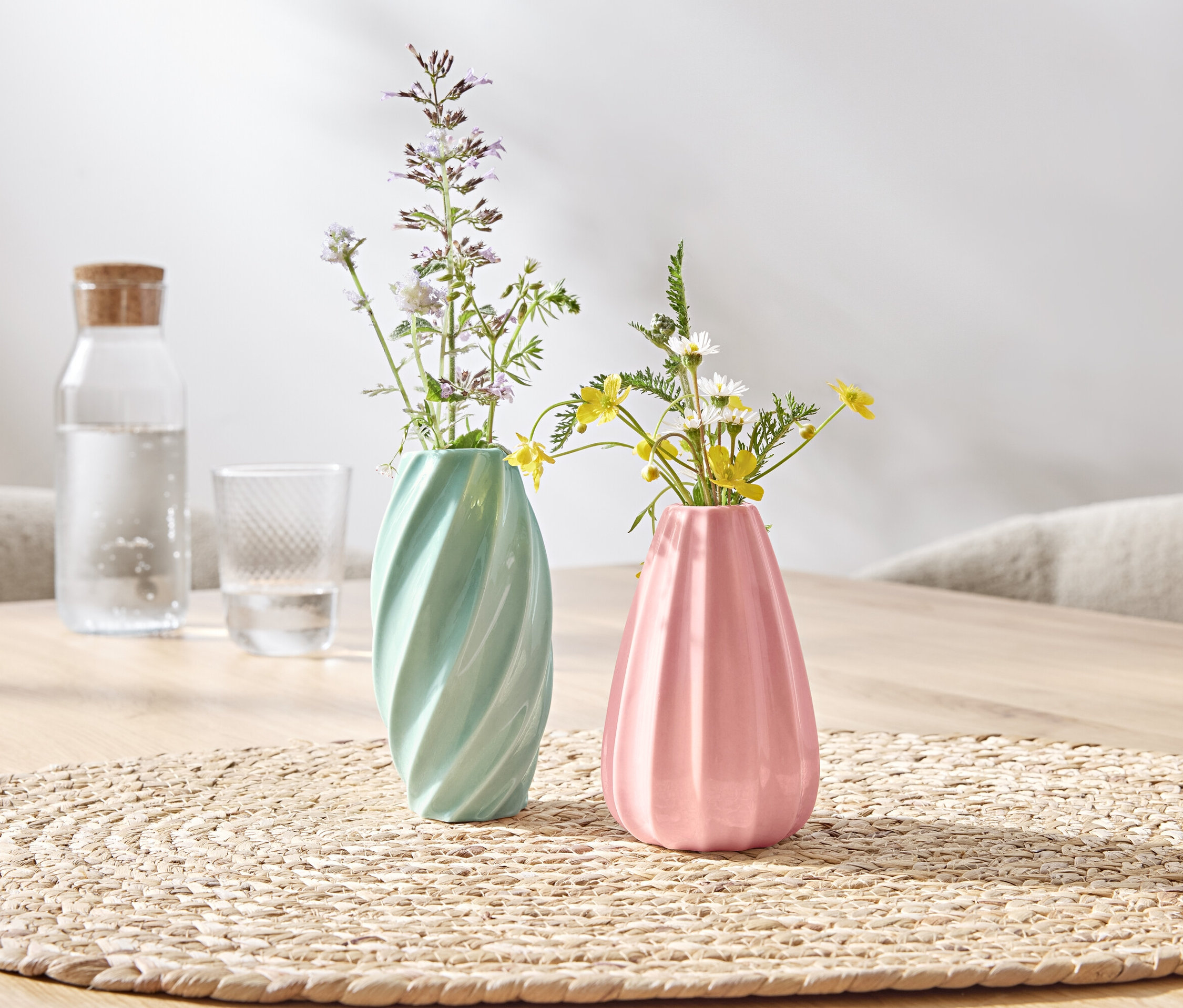 Sur une table, deux vases avec des fleurs, un vert et un rose.
