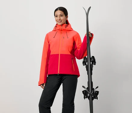Femme en veste rouge et orange tenant des skis.