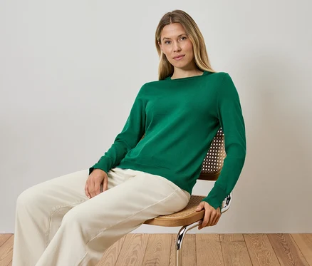 Femme blonde assise sur une chaise, portant un pull vert et un pantalon crème.
