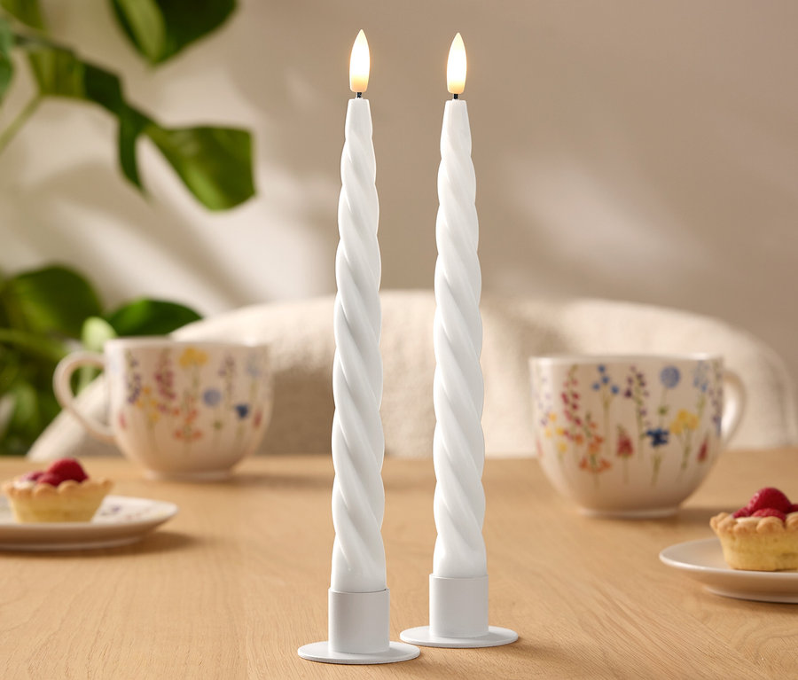 Deux bougies LED blanches en spirale sont posées sur une table en bois. À l'arrière-plan, on peut voir deux tasses à motif floral et de petites tartelettes.