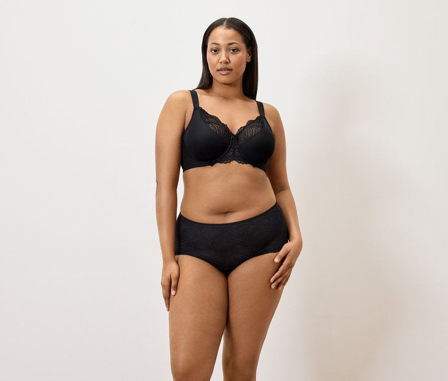Une femme porte un soutien-gorge minimiseur noir et 5 culottes noires sur un fond blanc.
