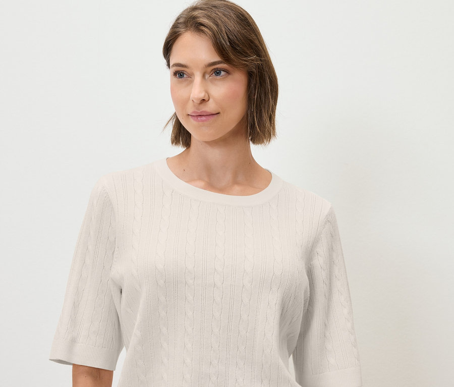 Une femme porte un pull en tricot torsadé crème à manches courtes.