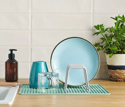 Une assiette bleue, un verre et un égouttoir avec tapis en silicone sur un comptoir de cuisine à côté d'un évier.