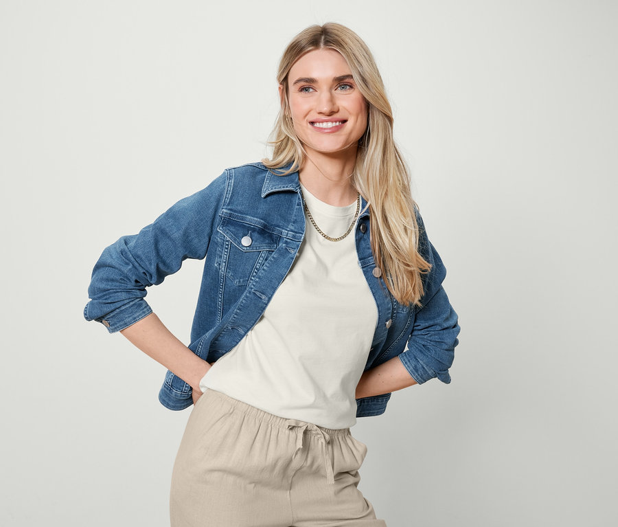 Une femme blonde souriante pose en veste en jean, t-shirt basique blanc et pantalon en lin couleur sable.