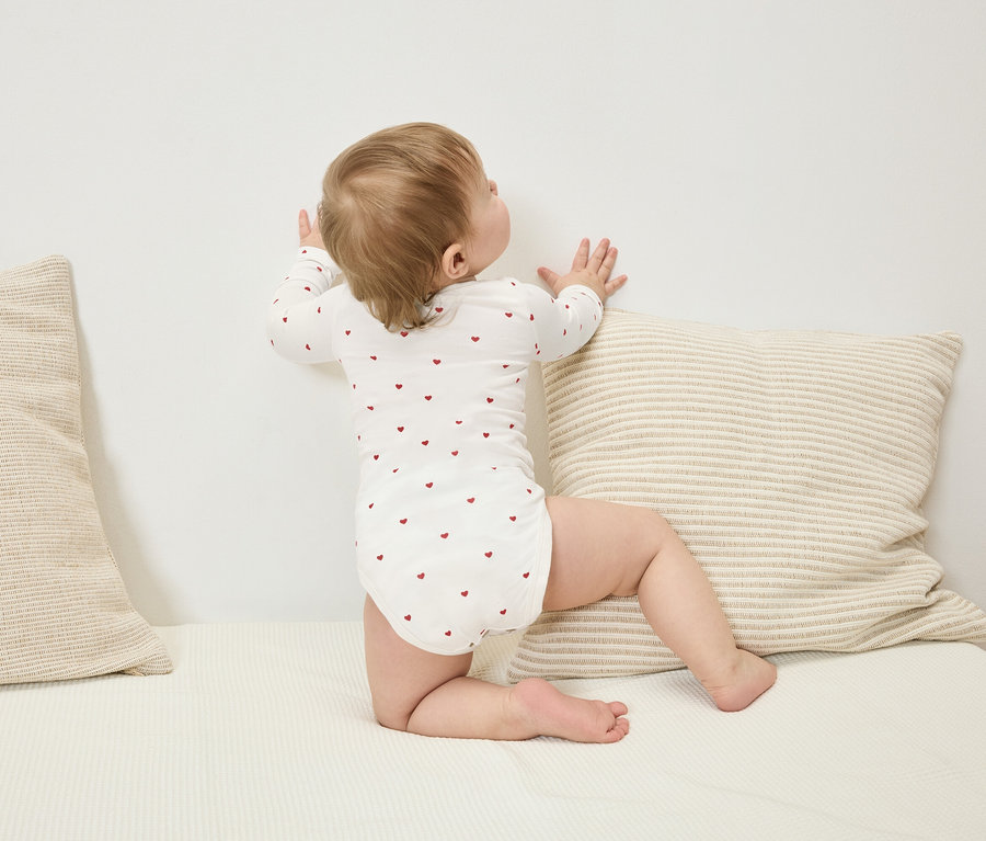 Un bébé en body à manches longues avec des cœurs rouges se tient contre un mur à côté d'un coussin rayé.