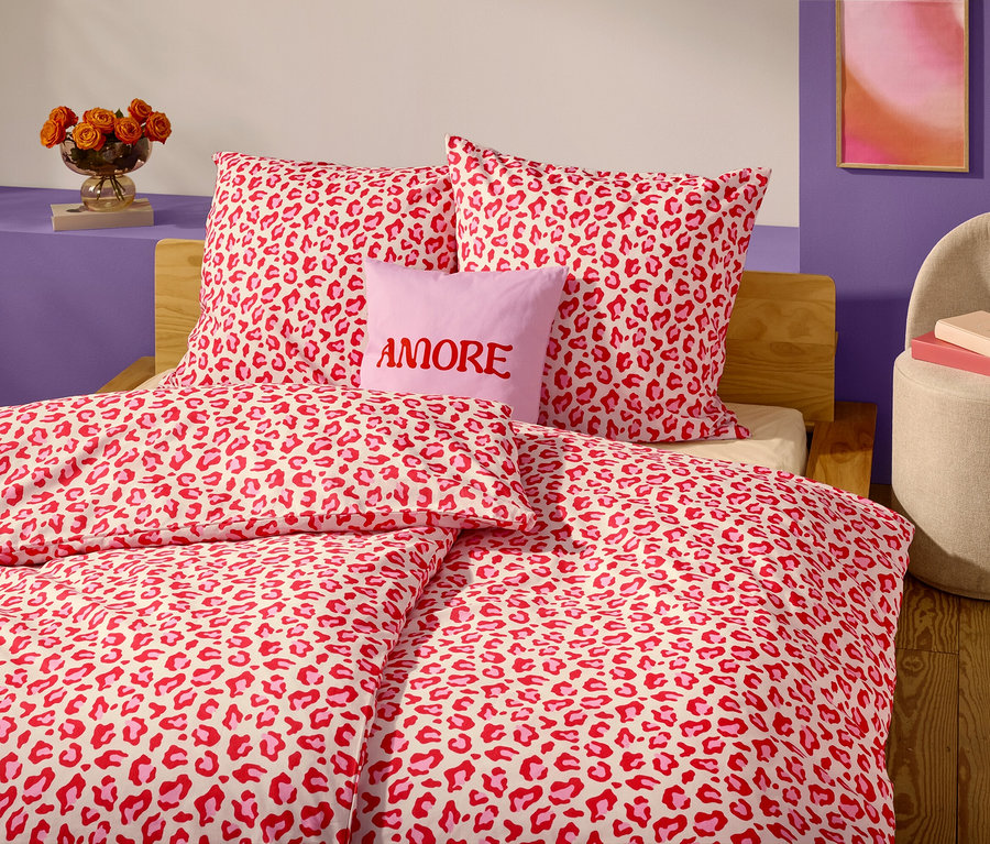Gros plan d'un lit avec des draps et des oreillers à motif léopard rouge et blanc. Un oreiller lavande avec le mot "AMORE" est placé devant deux oreillers.