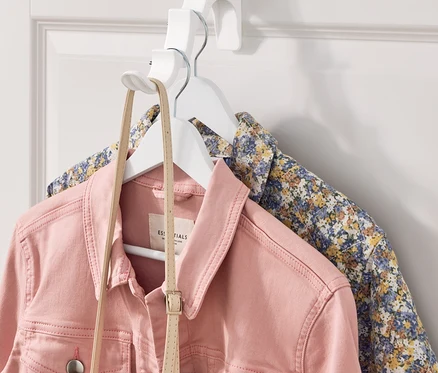 Une veste en jean rose et une chemise à fleurs sont suspendues à un cintre blanc fixé à un crochet de porte blanc.