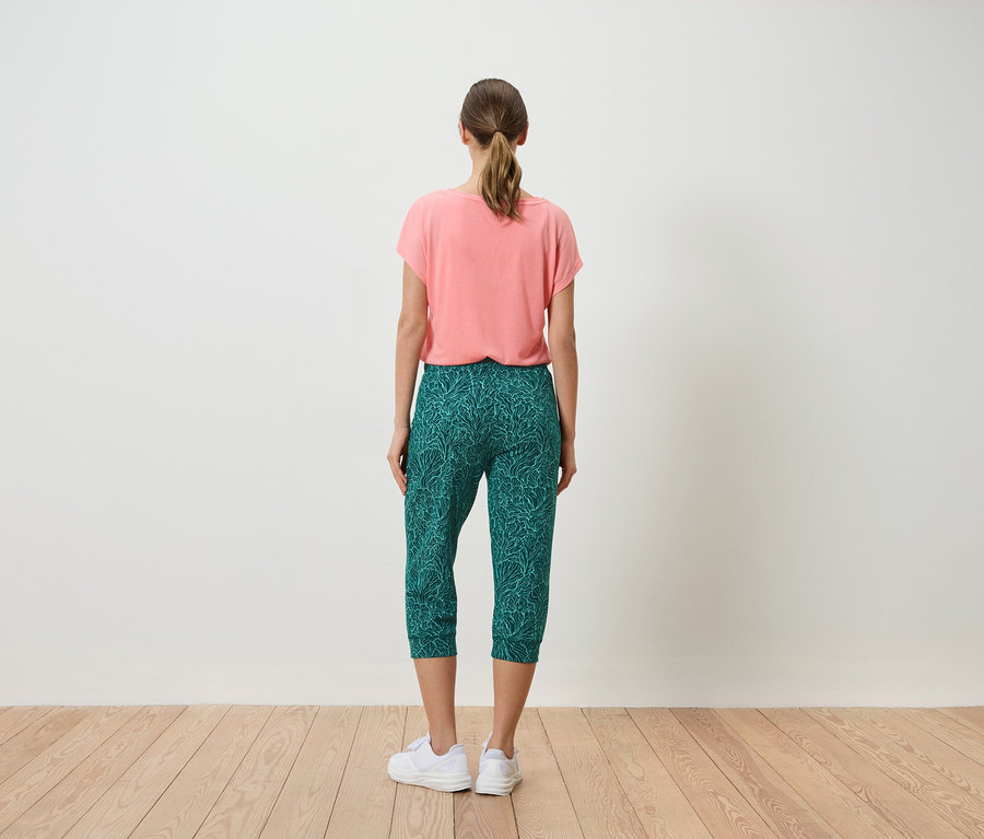 Femme vue de dos portant un t-shirt corail et un pantacourt vert avec un imprimé de feuilles, ainsi que des baskets blanches.