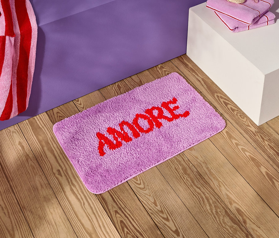 Un tapis de bain violet avec le mot rouge "AMORE" est posé sur un plancher en bois à côté d'une table de chevet violette.