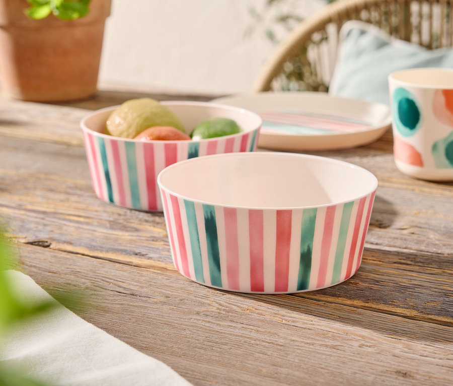 Sur une table en bois, deux bols rayés, dont un rempli de fruits, à côté d'une assiette, d'une tasse à pois colorés et d'une plante en pot.