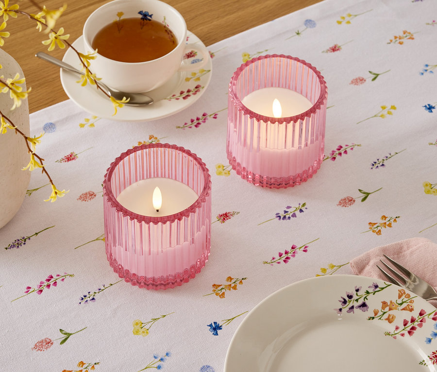 Sur une table à motif floral sont posés deux bougeoirs roses, une tasse de thé et une assiette.