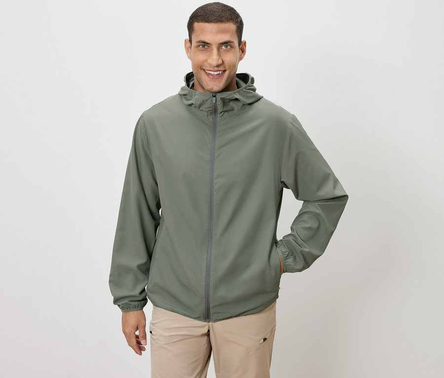 Homme portant une veste vert olive et un pantalon beige, une main dans la poche, sur un fond blanc.