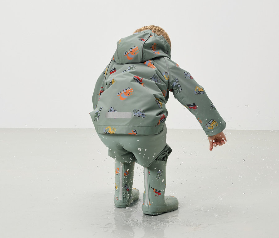 Un enfant vu de dos, portant un manteau de pluie vert et un pantalon de pluie avec un motif de pelleteuse et des bottes en caoutchouc assorties, jouant dans l'eau.