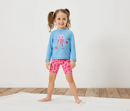 Une fillette avec des couettes porte un t-shirt bleu clair à manches longues avec une méduse rose et un short rose à motif d'écailles.
