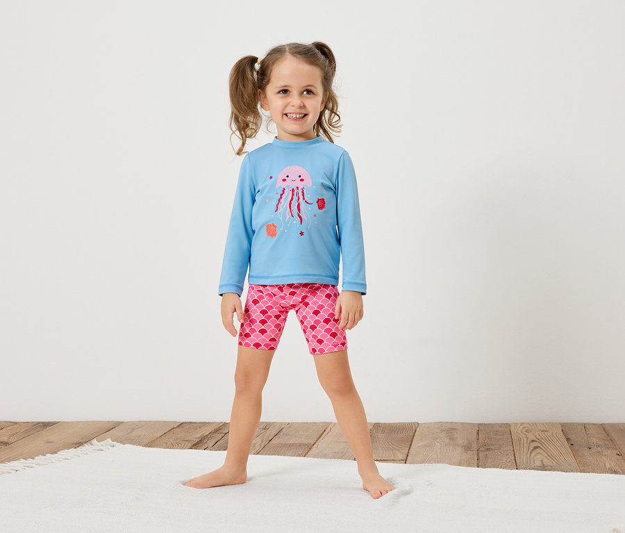Une fillette avec des couettes porte un t-shirt bleu clair à manches longues avec une méduse rose et un short rose à motif d'écailles.