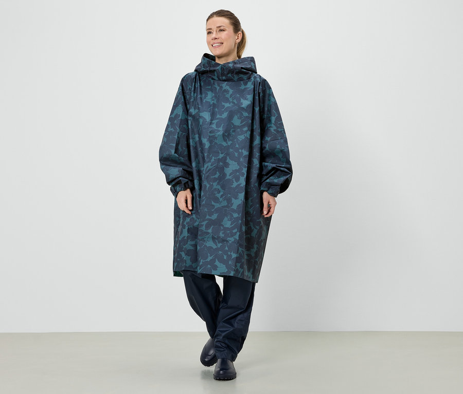 Une femme se tient debout dans un long poncho de pluie bleu à capuche avec un motif de feuilles.