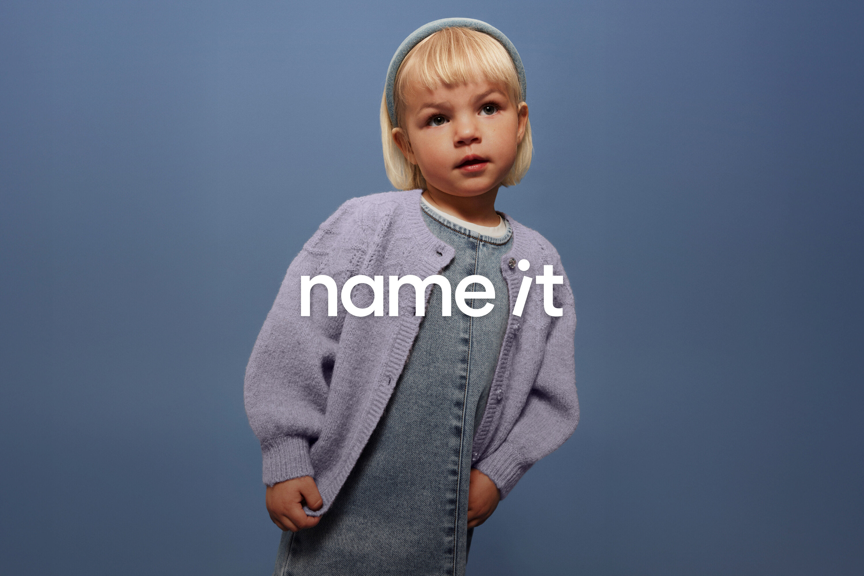 Un jeune enfant aux cheveux blonds et au bandeau bleu porte une salopette en jean et un cardigan lilas sur un fond bleu.
