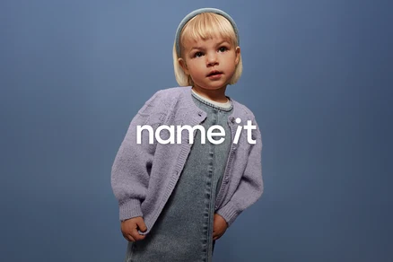 Un jeune enfant aux cheveux blonds et au bandeau bleu porte une salopette en jean et un cardigan lilas sur un fond bleu.