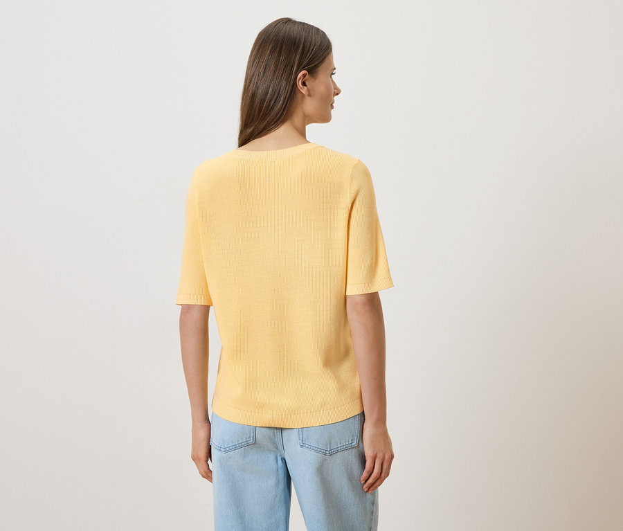 Une femme vue de dos, portant un jean bleu clair et un t-shirt jaune à manches courtes, sur un fond blanc.