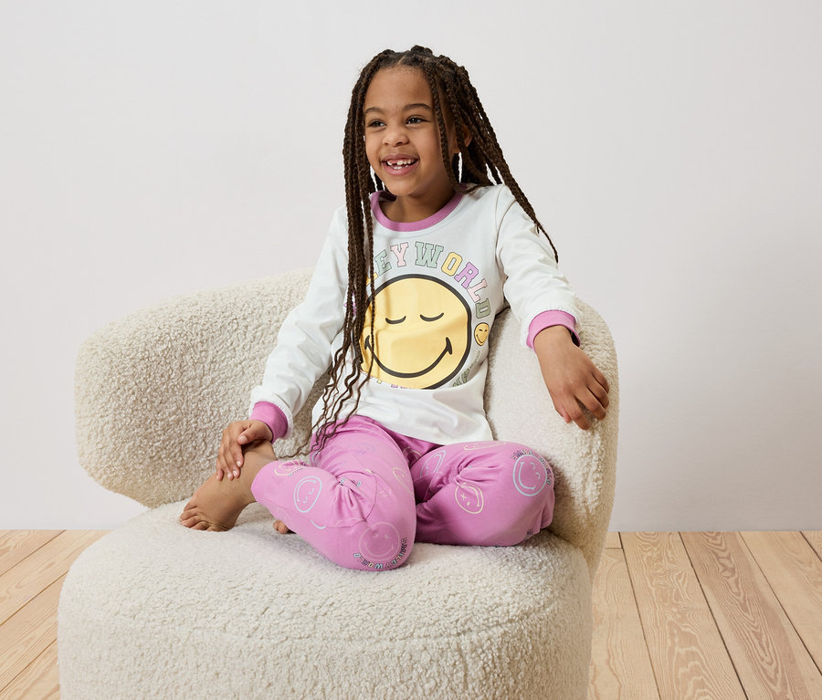 Une fillette souriante avec des tresses est assise pieds nus dans un fauteuil et porte un pyjama blanc avec un motif de smiley.