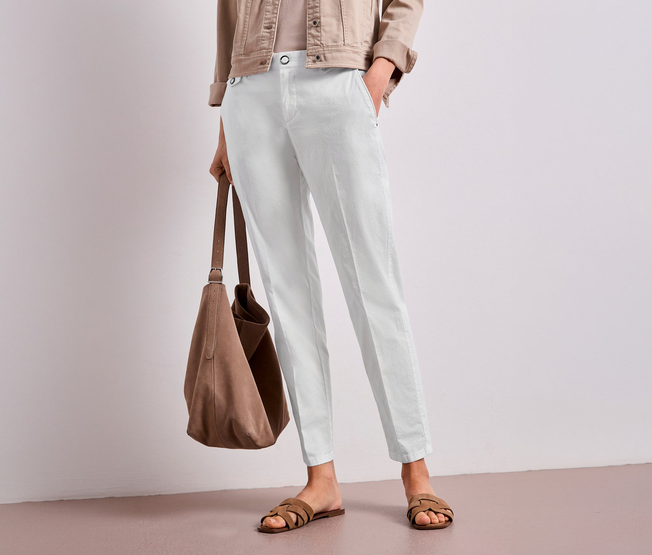 Une personne portant une veste marron clair, un haut clair et un pantalon chino Street One blanc est debout, tenant un sac en daim marron et portant des sandales marron. Une main est dans une poche du pantalon.