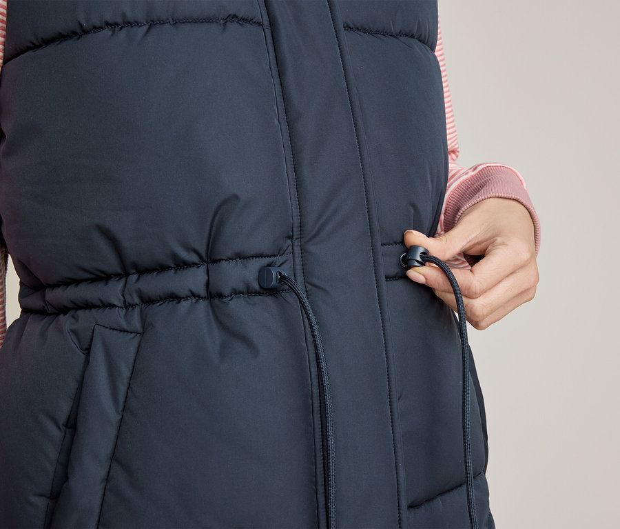 Gros plan d'une personne portant une doudoune sans manches bleu foncé sur une chemise rayée rose, ajustant le cordon de serrage à la taille du gilet avec sa main droite.