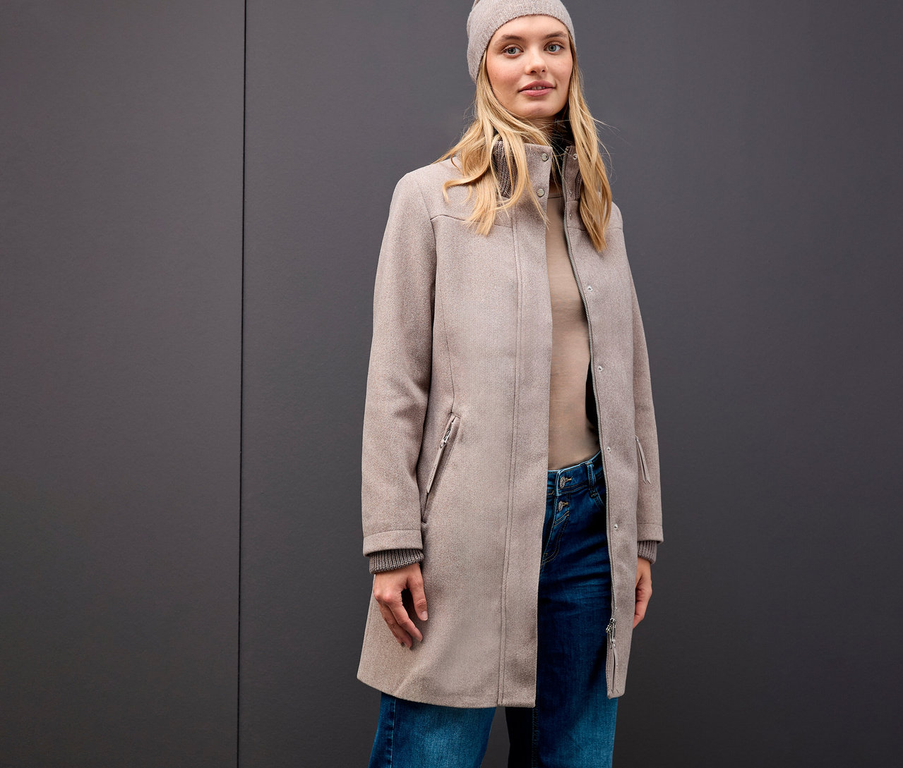 Une femme blonde portant un manteau court Street One, un bonnet, un col roulé et un jean devant un fond gris.